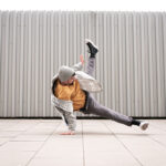 young white caucasian guy with beard gray clothes gray cap and yellow t-shirt dancing break dance doing acrobatics on the floor lifestyle gray metal background fence happy and contented